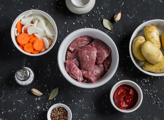 Raw ingredients - beef, potatoes, onions, carrots, tomato sauce on a dark stone background