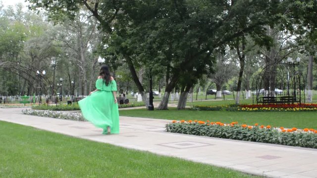 Счастливая довольная женщина брюнетка идет по аллее летнего парка в зеленом платье, кружится и позирует фотографу
