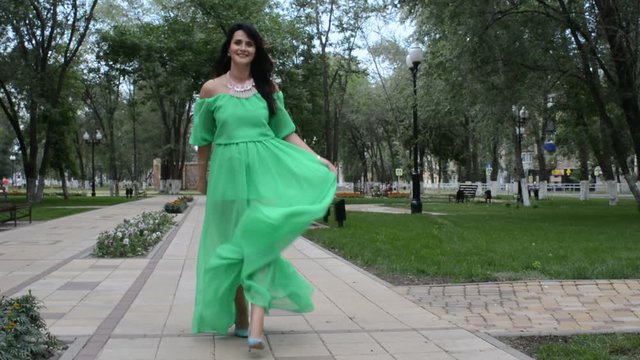Счастливая довольная женщина брюнетка идет по аллее летнего парка в зеленом платье, кружится и позирует фотографу
