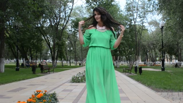 Счастливая довольная женщина брюнетка идет по аллее летнего парка в зеленом платье, кружится и позирует фотографу
