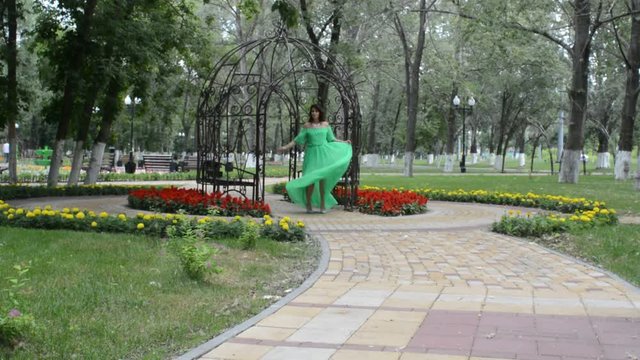 Счастливая довольная женщина брюнетка идет по аллее летнего парка в зеленом платье, кружится и позирует фотографу
