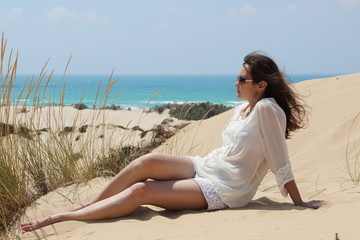 Fototapeta na wymiar Smiling brunette girl in sunglasses, a white blouse and jeans shorts is sitting on the sand on the background of the desert dunes and the sea