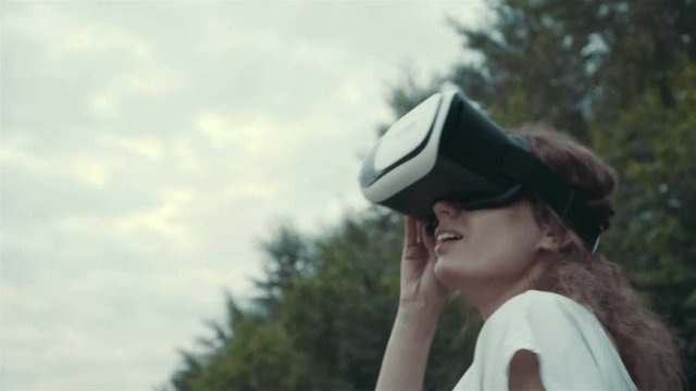 Young woman uses head-mounted display in the park. Playing game using VR-helmet for smart phones.