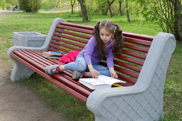 Cute little caucasian girl with beautiful long hair reading book sitting on a bench in spring park
