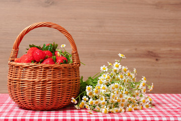 Fototapeta na wymiar Strawberries in a wicker basket and a bouquet of field chamomiles on a wooden background. Red checkered tablecloth.