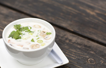 Thai soup in a white dish on a wooden table. Spicy coconut milk soup with shrimp and greens. 