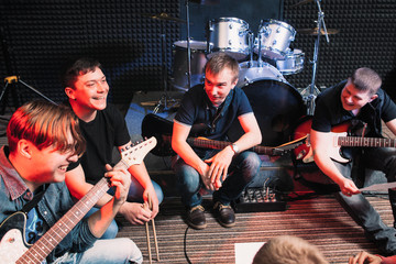 Laughing happy musicians sitting with instruments at studio. Guitarists and drummer discussing some...