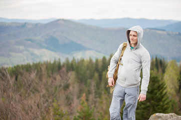 Fototapeta na wymiar Young tourist in a jacket with a hood with a backpack on his shoulder looking away, against forest and mountains in background