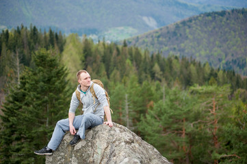 Naklejka na ściany i meble Athlete man with a brown backpack sitting on the peak of rock and looking down on the blurred background of forest valley and hills. Spring nature