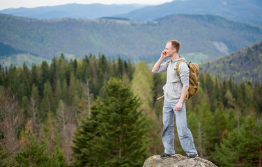 Fototapeta na wymiar Young male climber with backpack standing on top of a mountain looking away and talking on the phone on the blurred background of forest valley and hills