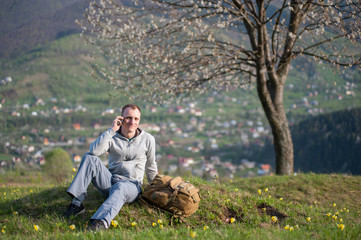 Naklejka na ściany i meble Traveler with backpack sitting on hill with green grass and talking on the mobile phone on the blurred background of blooming tree and small village in the distance