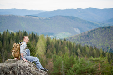 Naklejka na ściany i meble Young tourist man with a backpack sitting on the edge of a rock with an astonishing blurred view on forest valley and hills