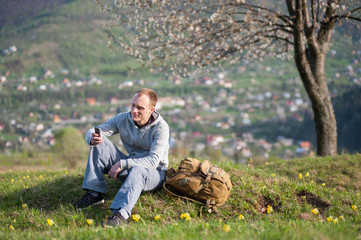 Naklejka na ściany i meble Traveler male with backpack sitting and holding a mobile phone, on hill with yellow flowers near lonely blooming tree with small town and forest valleys as background.