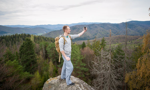 Young courageous hiker with brown backpack standing on the peak of a rock and makes selfie on the his phone with forest valley and nice hills. Wide angle lens