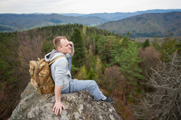 Male backpacker with a backpack is relaxing on the top of rock, looking away and talking on the phone on the background of forest valley and hills.
