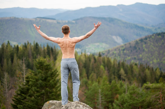 Young man with a bare torso standing on the peak of a rock, with back to the camera with hands up, against picturesque view of on the green forest and nice mountains