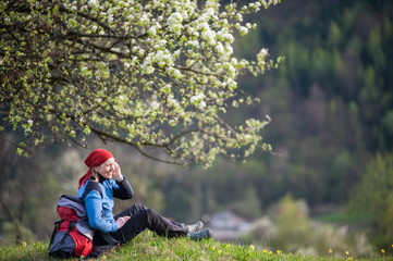 Fototapeta na wymiar Happy female with a backpack relax under blooming tree on top of hill with yellow wildflowers, green grass and talking on the mobile phone with forest valleys as blurred background.