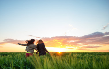 two girl friends running into wheat to the sunset - obrazy, fototapety, plakaty