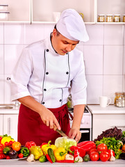 Man in chef hat keeps knife and cooking fresh vegetable salad. There are vegetable on foreground. Vegetarian cooking.