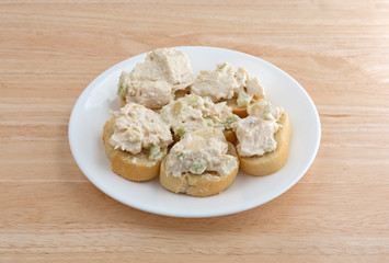 Chicken salad on small pieces of French bread with white plate atop a wood table.