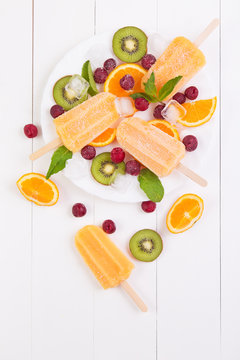 Homemade orange popsicles on a plate with mint, kiwi, frozen cherry and orange slices on a plate and white wooden background. Top view with copyspace