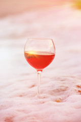 glass with pink wine in a white sea foam on the beach.  In the background, emerald wave rolls ashore. Natural beautiful backdrop and sea glass
