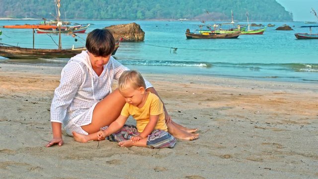 Baby and mother draw in the sand with a stick on the beach with boats in the sea in background