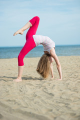 Fototapeta premium Young lady practicing yoga. Beautiful woman posing at the summer sand beach. Workout near ocean sea coast. Beautiful fit tan girl. Fitness model caucasian ethnicity outdoors. Weight loss exercise