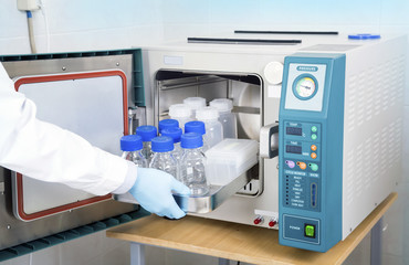 Scientific researcher hand in blue glove inserts tray with glass and plastic bottles into the...
