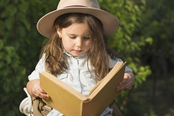 Adorable little girl in a safari hat and explorer clothes reading old book sitting in a wooden suitcase with backpack and safty roap outdoor