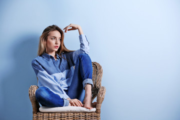 Beautiful young woman in shirt and jeans sitting on chair on blue wall background