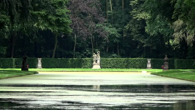 Romantic small lake with statues on the side pond located at green silent quiet scenery trees foliage branches all around it  on both sides of small pond green surroundings of lake quiet water 4k