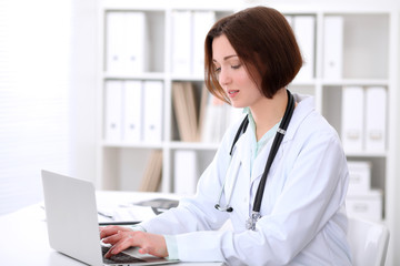 Young brunette female doctor sitting at the table and typing on laptop computer.    Health care, insurance and help concept. Physician ready to examine patient