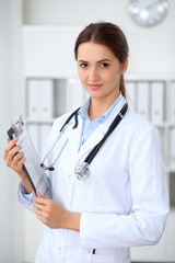 Young brunette female doctor standing with clipboard and smiling at hospital.  Physician ready to examine patient