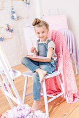 Little artist in denim overalls painting brushes on the easel in her studio