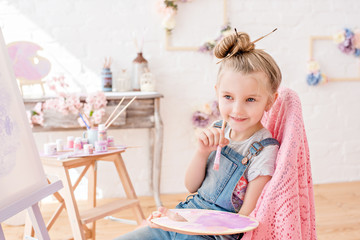 Fototapeta premium Little artist in denim overalls painting brushes on the easel in her studio