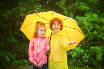 Brother and sister stand together under a big yellow umbrella.