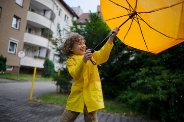 Cheery lad walks in the rain in the yard of the house with a yellow umbrella.