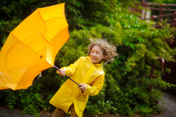 The boy in a bright yellow raincoat with effort holds an umbrella from wind.