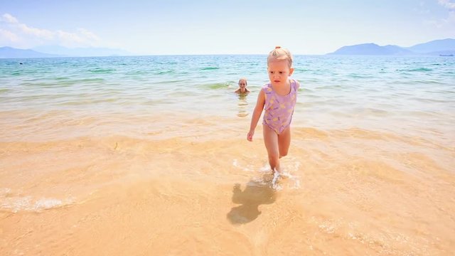 Blond Little Girl Walks out of Sea to Sand Castle on Beach