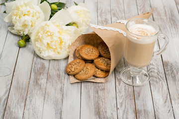 Glass of cappuccino next to cookies and pionies flowers on white wooden background. Place for text.