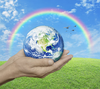 Earth in hands over green grass with blue sky, cloud, rainbow an