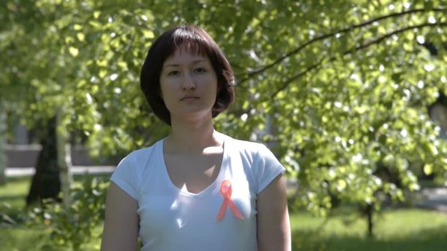 Young brunnete woman in white t-shirt wearing orange ribbon, trees background. Leukemia, melanoma, multiple sclerosis, Self Injury Awareness Day
