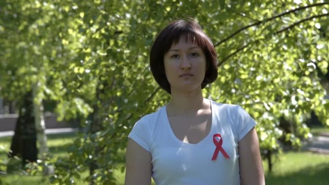 Young brunnete woman in white t-shirt wearing red ribbon, trees background. AIDS and HIV awareness, heart disease, lymphoma, tuberculosis, substance-abuse, vasculitis
