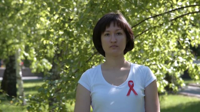 Tilt shot of young woman in white t-shirt with red ribbon, green nature background. AIDS and HIV awareness, heart disease, lymphoma, tuberculosis, substance-abuse, vasculitis