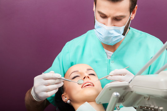 Male dentist examines the mouth of the young beautiful woman patient with perfect straight white teeth on the dentist's chair with the help of dental mirror. Healthcare, medicine.