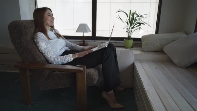 Attractive female has video call on the computer. Wife happy and smiling tell emotionally stopy with friendly smile. She sitting on the armchair near window wearing in white shirt and shoes with high