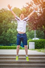 Fototapeta premium Happy jogger jumping on the stairs in the park with raised hands. Another successful training is finished. Sport and fitness success concept. 