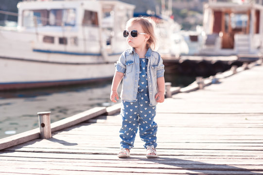 Stylish baby girl wearing denim suit and jacket at wooden pier. Summer season. Child standing outdoors.
