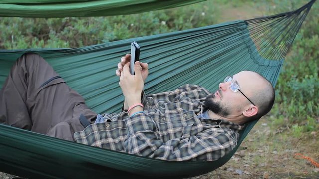 A man in a hammock is video calling on the phone. Man in the woods with a beard and glasses
